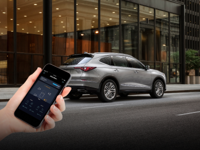 Shot of a hand holding a smart phone displaying the AcuraLink™ app in the foreground of an image. The background image features a grey Acura MDX parked in front of a futuristic building./Prise de vue d'une main tenant un téléphone intelligent affichant l'application AcuraLinkMC au premier plan d'une image. L'image de fond présente une Acura MDX grise stationnée devant un bâtiment futuriste.