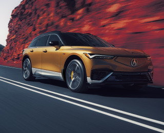 3/4 bird’s eye front view of gold ZDX driving on mountain highway. Part of the image is saturated with colour, so the dirt on the sides of the highway is red.